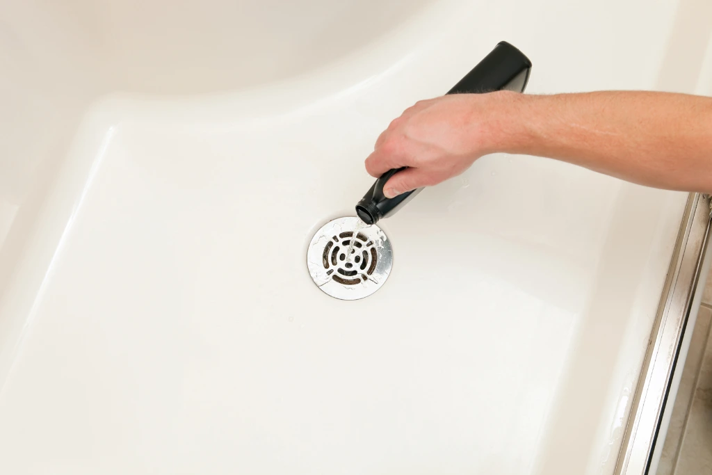 A hand pours liquid drain cleaner from a black bottle into a round, stainless steel bathtub drain. The white surface of the bathtub surrounds the drain.

