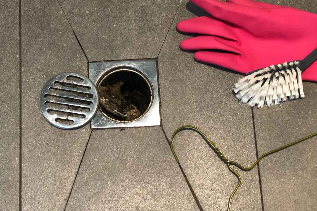 Close-up of a floor drain clogged with hair and debris. The metal grate has been removed, and cleaning tools, including a pink glove and brush, are positioned next to it.

