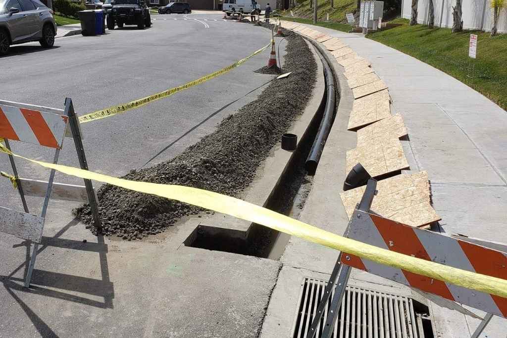Drains Pros Ventura repairs an area drain in Ventura County. A trench with pipes and gravel, caution tape, and safety barriers mark the work zone. A grey car is nearby, with residential buildings in the background.