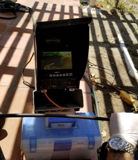 A Drains Pros Ventura technician uses a drain inspection camera with a monitor displaying an internal pipe view. The camera system sits on a blue toolbox, with a white PVC pipe and yellow floating device visible in the background, all under striped shadows from a patio cover.