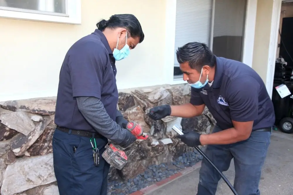 Two Drains Pros Ventura technicians, wearing blue shirts and face masks, are performing drain cleaning services. One technician holds a red power tool while the other feeds a black cable into a drain opening near a rock-faced wall.
